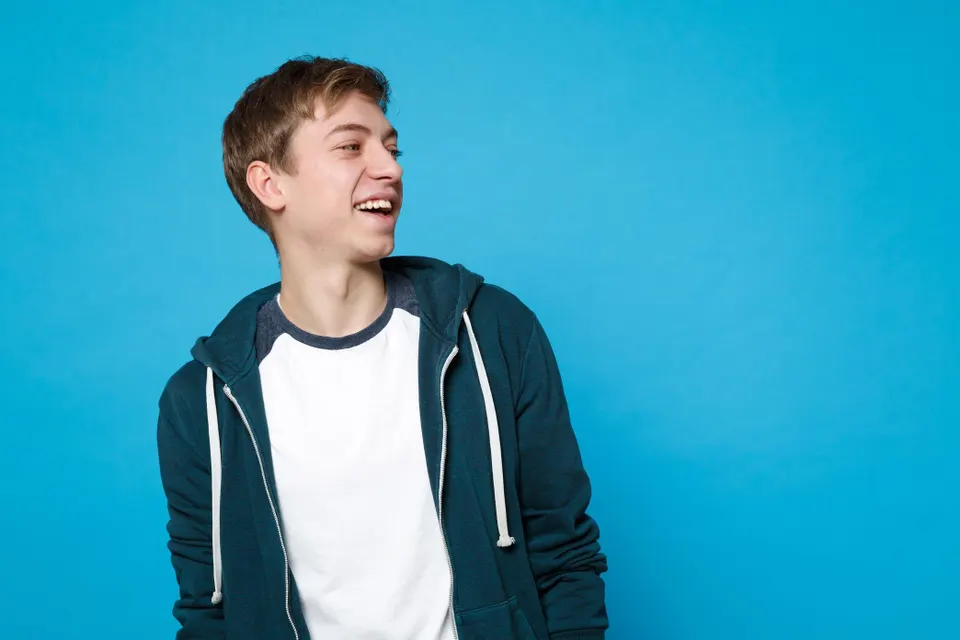 Young man laughing against a bright blue background, wearing a teal hoodie over a white T-shirt.
